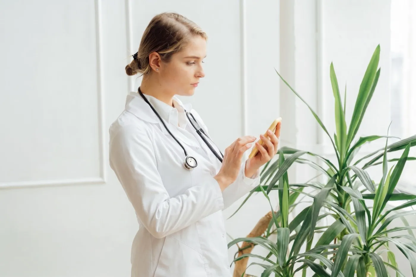 Doctora con bata blanca revisando su WhatsApp junto a una planta en la clínica